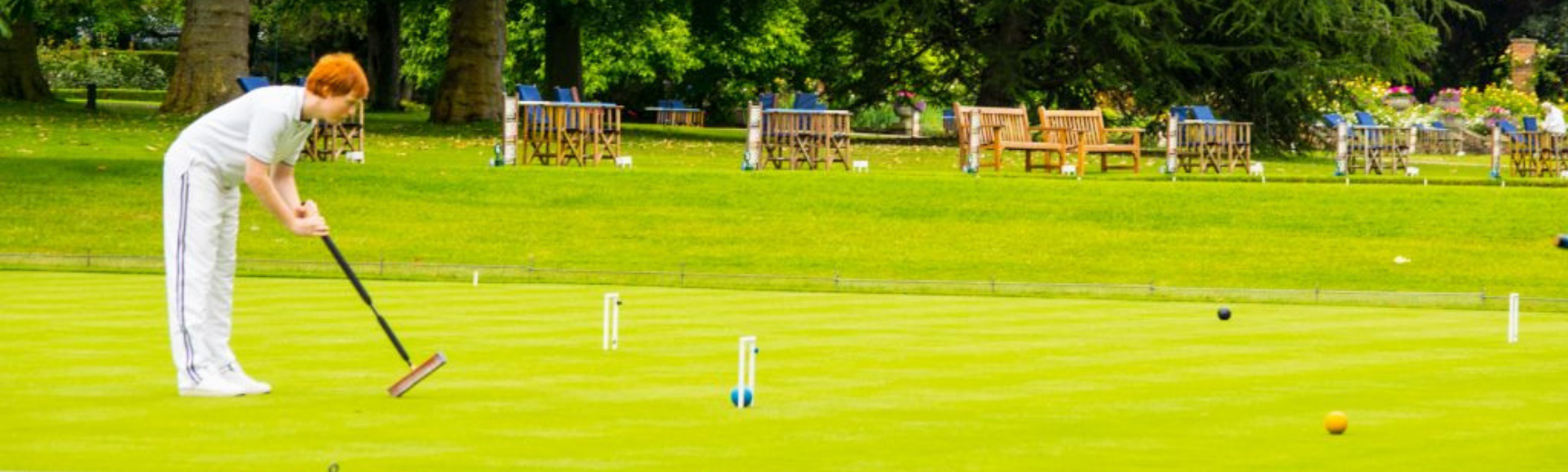 A man in all white hitting a croquet ball with a mallet, across a croquet lawn