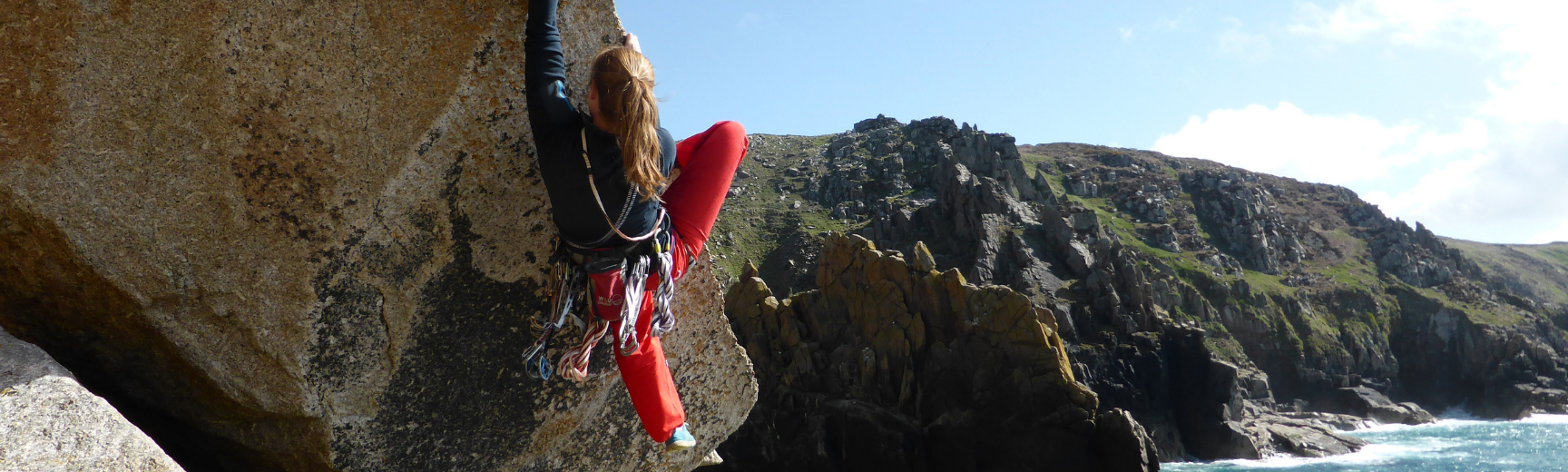 A climber climbing up an angled rock, with a view of the coastline behind her