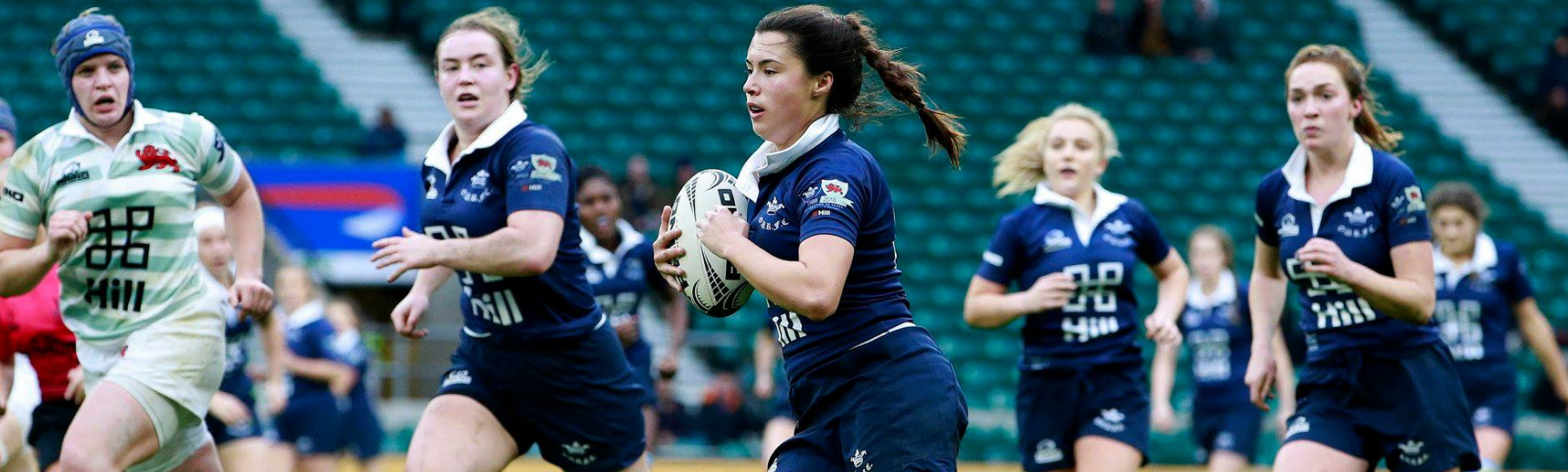 A women in dark blue carrying a rugby ball in the Varsity match against Cambridge, supported by several teammates, at Twickenham Stadium