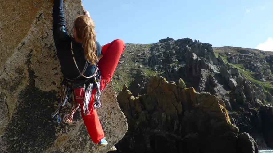 A climber climbing up an angled rock, with a view of the coastline behind her