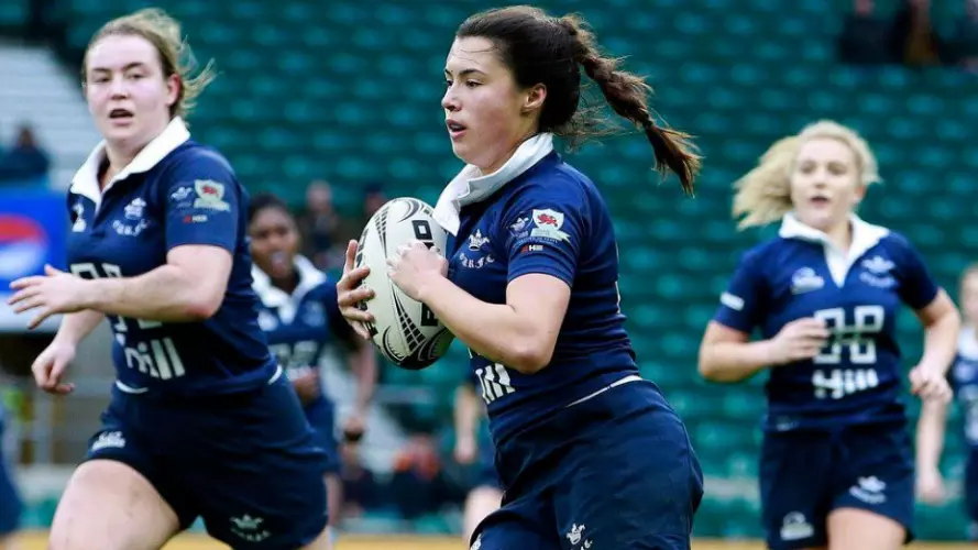 A women in dark blue carrying a rugby ball in the Varsity match against Cambridge, supported by several teammates, at Twickenham Stadium